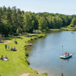 Vue d'ensemble ensoleillée du plan d'eau, de la forêt environnante et des familles profitant des activités au Parc Loisirs et Nature de la Porte du Hainaut.